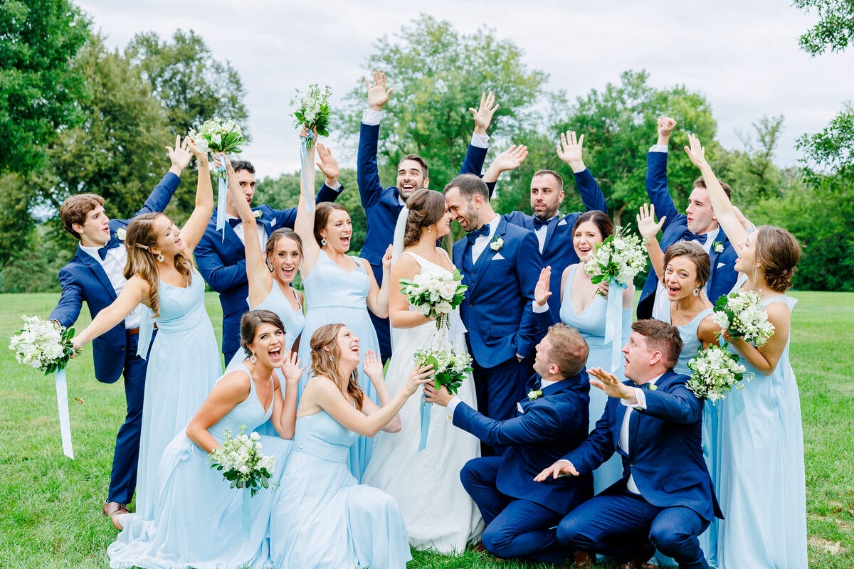 Wedding party cheers around the bride and groom. Bridesmaids wearing light blue dresses and groomsmen wearing navy blue suits. Wedding party photos in a park, Sioux Falls, South Dakota